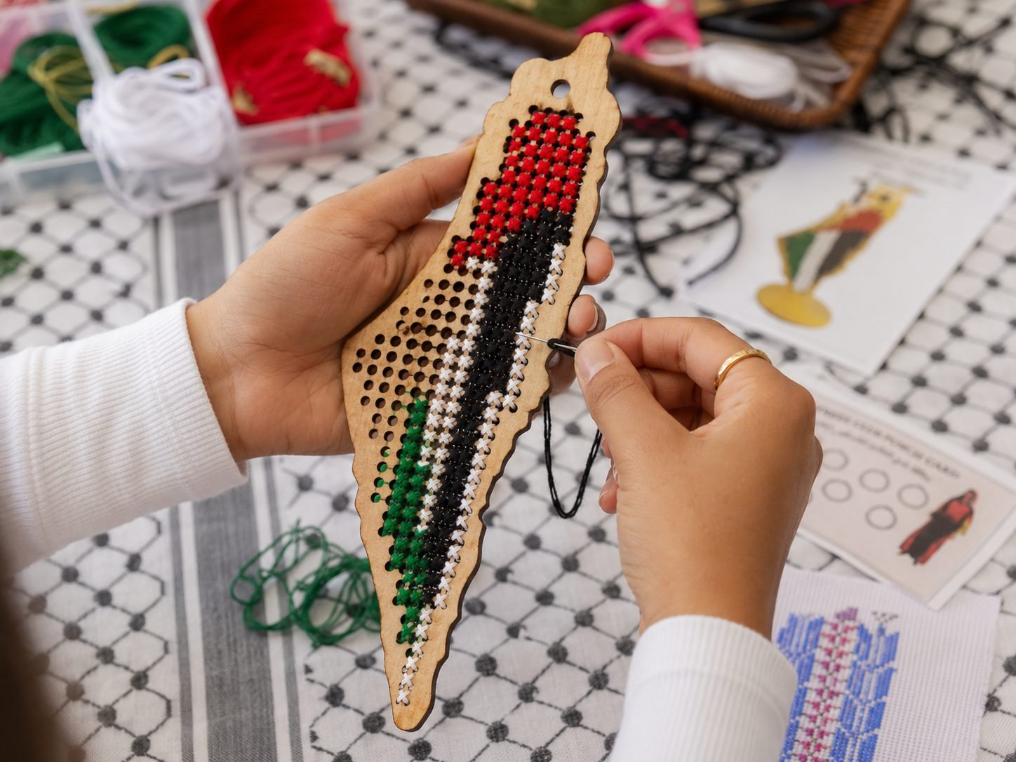 Hands stitching a tatreez pattern on a wooden Palestine-shaped board